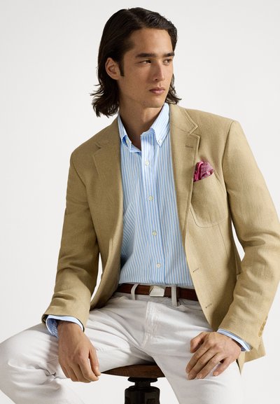 Ανοιχτό μπεζ σακάκι πάνω από μπλε και λευκό ριγέ πουκάμισο με κουμπιά, με ροζ μαντήλι στην τσέπη. Λευκό παντελόνι και καφέ ζώνη ολοκληρώνουν το σύνολο.