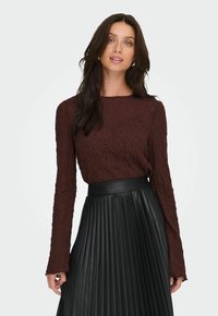 Donna con lunghi capelli scuri che indossa un top a maniche lunghe texturizzato color amaranto e una gonna nera plissettata a vita alta, su uno sfondo neutro.