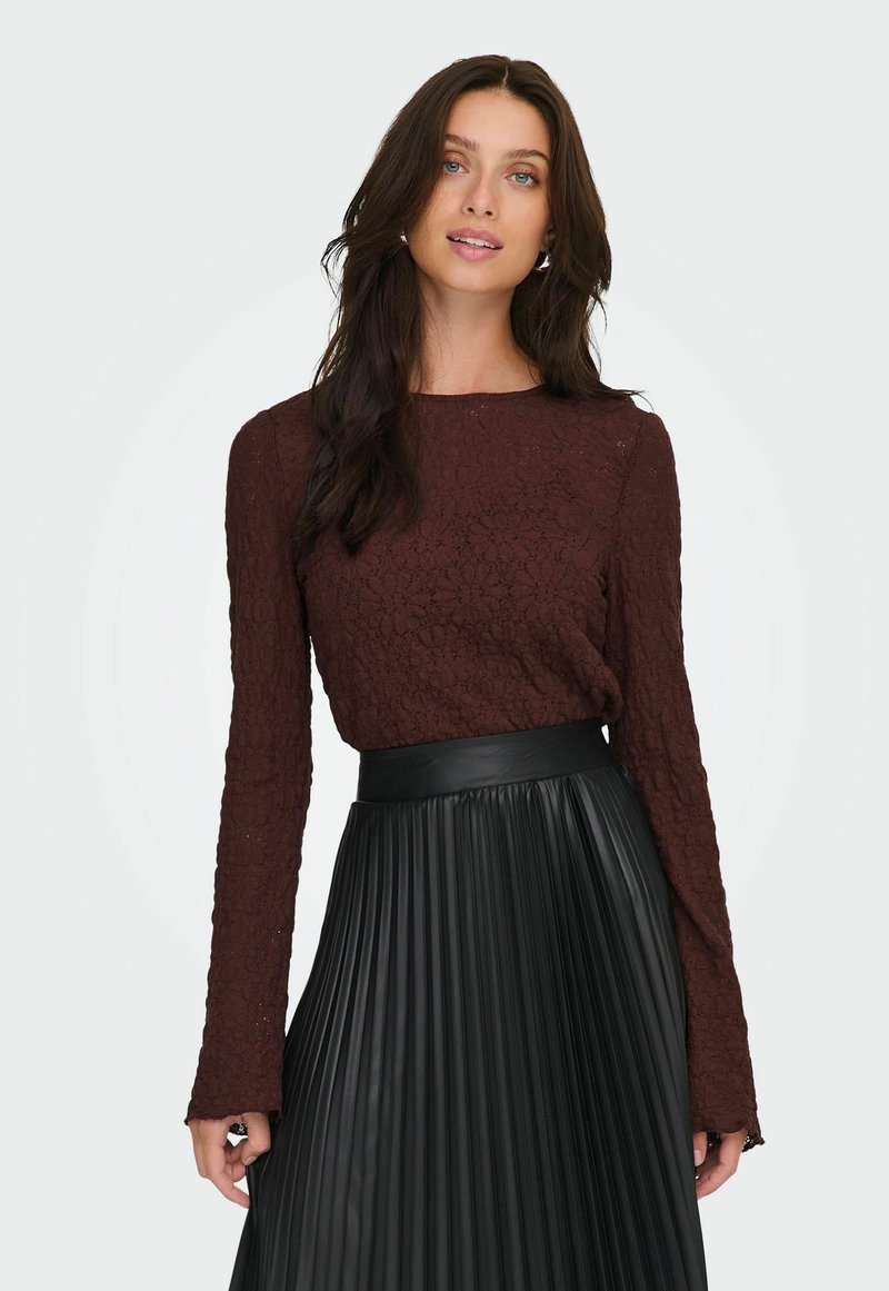 Donna con lunghi capelli scuri che indossa un top a maniche lunghe texturizzato color amaranto e una gonna nera plissettata a vita alta, su uno sfondo neutro.