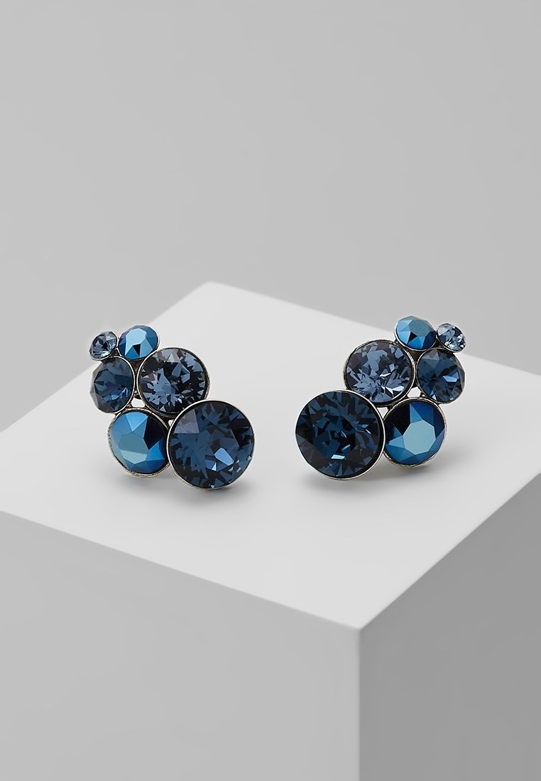 Groupe de boucles d'oreilles en pierre précieuse facettée bleu foncé, disposées en tailles variées sur un piédestal géométrique blanc sur un fond gris.