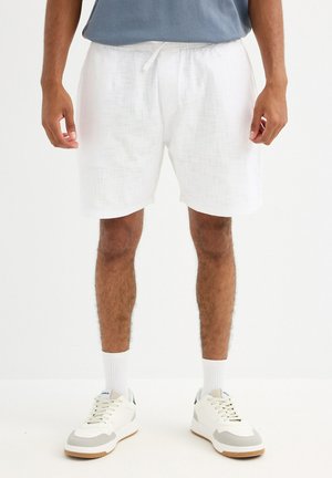 Hombre con pantalones cortos blancos con cordón, calcetines blancos hasta el tobillo y zapatillas blancas, posando frente a un fondo liso.