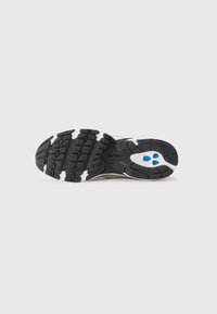 Semelle en caoutchouc noire d'une basket avec des motifs de la bande de roulement et trois détails circulaires bleus près du talon sur un fond gris clair.