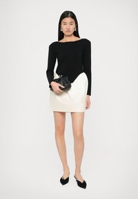 Μαύρη μπλούζα με μακριά μανίκια και φαρδιά λαιμόκοψη, συνδυασμένη με μια ασυμμετρική λευκή φούστα. Το μοντέλο κρατάει μια μαύρη τσάντα clutch και φοράει μαύρα ψηλοτάκουνα παπούτσια.