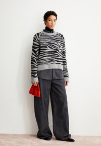 Grauer Pullover mit schwarzen Zebra-Streifen, grauen Bündchen und Kragen, kombiniert mit lockeren schwarzen Jeans. Das Model hält eine rote, gerippte Mütze.