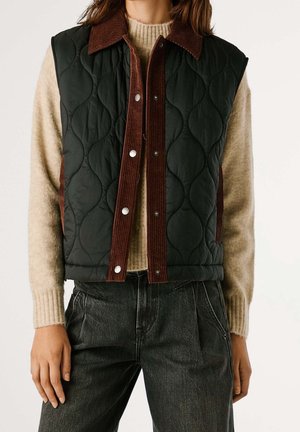 Person trägt eine schwarze gesteppte Weste mit braunem Cordbesatz über einem beigen Pullover und dunklen Jeans, steht vor einem schlichten Hintergrund.