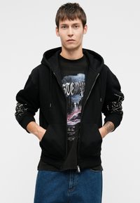 Jauns vīrietis ar rokām kabatās, uzvilcis melnu kapuci ar baltu piedurkņu dizainu, grafisku t-kreklu un zilo džinsu, fona priekšā bez apdrukas.