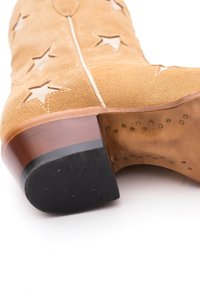 Stivale cowboy in suede beige con fori a forma di stella e cuciture bianche. Tacco in legno con base in gomma nera e texture a pois. Interno in pelle liscia.