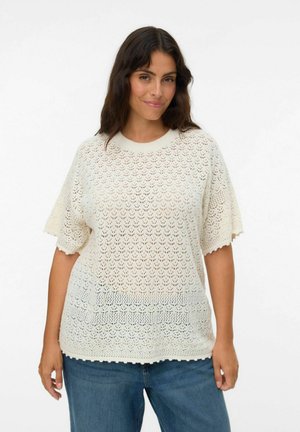 Pull en maille crème léger, à manches courtes, avec un motif en trame lâche et des bords festonnés. Associé à un jean en denim bleu.