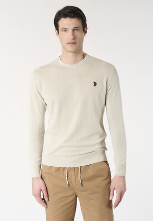 Uomo che indossa un maglione beige girocollo con piccolo logo e pantaloni beige con coulisse, in piedi davanti a uno sfondo chiaro uniforme.