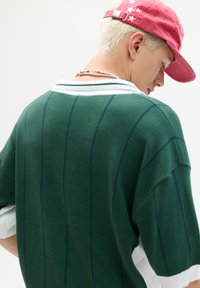 Grøn oversized sweater med lodrette striber, hvid krave og kontrasterende manchetter. Modellen har en rød hat og lagdelte sølvhalskæder på.
