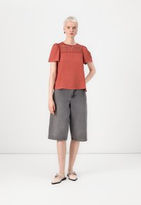 Top color ruggine con dettagli in pizzo sullo scollo, maniche corte a sbuffo; abbinato a pantaloni bermuda in denim grigi e ballerine beige.