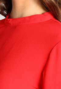 Röd topp med en texturerad tygkvalitet, rund halsringning och korta ärmar, som visar en slät, livlig färg utan utsmyckningar.