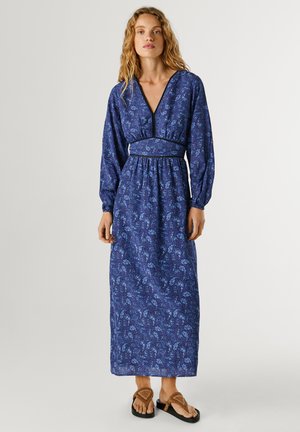 Vrouw draagt een lange blauwe jurk met patroon, met lange mouwen en een V-hals, gecombineerd met bruine sandalen, staand tegen een effen achtergrond.