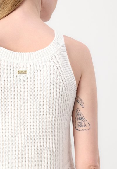 Débardeur en tricot blanc avec une texture côtelée, des bretelles larges et une petite étiquette en logo doré à l'arrière. Un tatouage de couleur peau visible sur le bras.