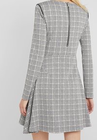 Mujer vestida con un vestido de patrón de pata de gallo en blanco y negro, de manga larga, con cremallera trasera y falda acampanada, vista desde la espalda.