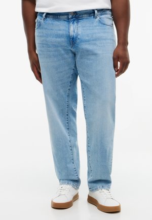 Mand iført lyseblå denimjeans og hvide sneakers med brune såler står mod en hvid baggrund.