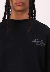 Persona con capelli intrecciati che indossa una maglietta nera a costine con la scritta "New Era Since 1920" ricamata in grigio sul petto.