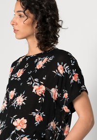 Blusa preta com padrão floral e mangas curtas, apresentando um tecido texturizado, padrões de rosas em tons de rosa e azul, e um corte descontraído com decote arredondado.