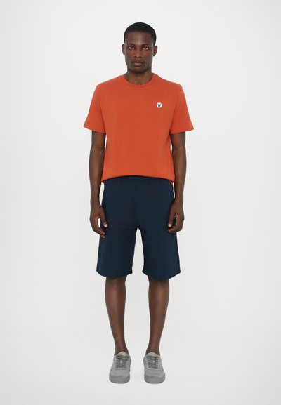 Orange short-sleeve t-shirt with a small logo, paired with navy shorts. Gray sneakers complete the outfit against a plain white background.