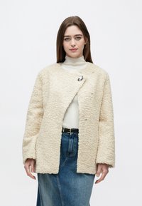 Vrouw draagt een crèmekleurig gestructureerd jasje over een witte coltrui en een blauwe denim rok met een zwarte riem, staand tegen een witte achtergrond.