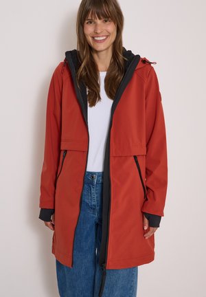 Donna sorridente che indossa una lunga giacca rossa con cappuccio sopra una camicia bianca e jeans blu, in piedi davanti a uno sfondo chiaro uniforme.