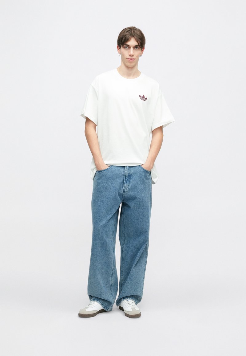 Witte oversized T-shirt met een bordeaux logo, gecombineerd met lichtblauwe wijde jeans en witte sneakers met bruine accenten.