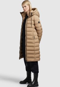 Beiger, gepolsterter Parka mit Kapuze, ausgestattet mit einem Reißverschluss, Seitentaschen und einem verstellbaren Kordelzug. Glänzende, glatte Textur. Schwarze Stiefel.