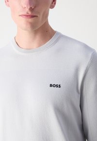 Svetlo sivi pulover z teksturirano zgornjo sekcijo, rebrastim ovratnikom in majhnim črnim logotipom "BOSS" na prsih. Mehka tekstura tkanine.