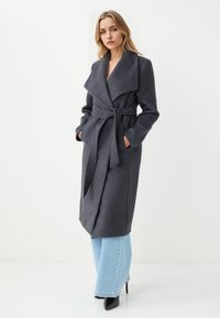 Grijze wikkelcoat met een brede kraag, een ceintuur in de taille en zijzakken, gecombineerd met lichtblauwe wijde jeans en zwarte puntige hakken.