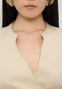 Gros plan sur le cou et les lèvres d’une femme portant un haut beige à col en V et de grandes boucles d’oreilles en or avec de longs cheveux foncés.