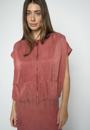 Noor pruuni pikkade juustega noor naine, kes kannab tolm-roosat varrukateta lühikest lukuga pluusi ja sobivat seelikut, seisab lihtsa tausta ees.