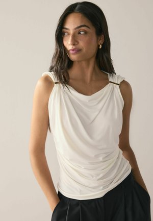 White draped top with soft texture, featuring gathered details at shoulders and gold-tone accents, paired with black high-waisted pants.