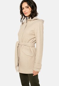 Manteau en mélange de laine beige avec capuche, taille ceinturée et poches latérales. Présente une finition texturée et des accents de boutons sur les poches.