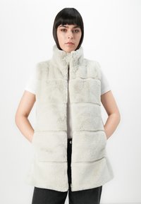 Nepbont vest in lichtgrijs, met een hoge kraag, horizontaal gewatteerd ontwerp en zijzakken. G gedragen over een wit shirt met een zwarte broek.