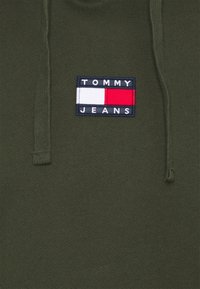 Oliveová zelená mikina s kapucí s texturovaným povrchem, se čtvercovou nášivkou loga Tommy Jeans v námořnické modři, bílé a červené. Detaily s tkaničkami na krku.