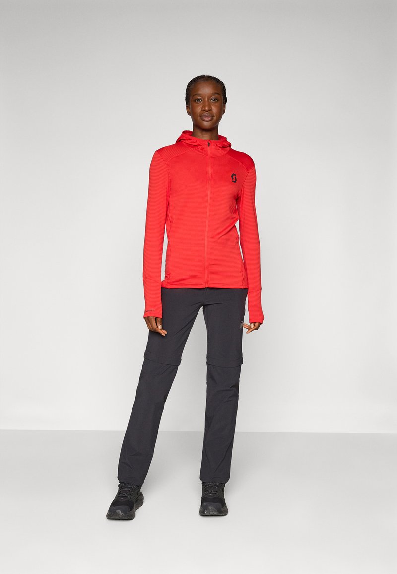 Czerwona bluza z kapturem na zamek błyskawiczny z teksturowanym wzorem i logo, zestawiona z czarnymi spodniami z odpinanymi nogawkami. Całość dopełniają ciemne obuwie.