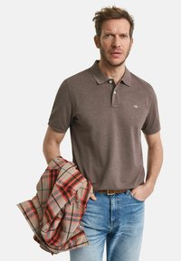 Brun polo skjorta med två-knappars knäppning, bärs med ljusblå denimjeans. Han håller en röd och beige rutig flanellskjorta.