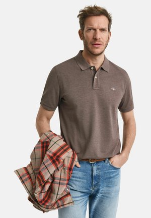Braunes Polohemd mit двухknopf-manager, getragen mit hellblauen Jeans. Er hält ein rot-beige kariertes Flanellhemd.