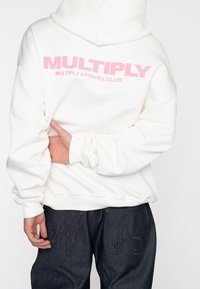 Witte hoodie met roze "MULTIPLY" tekst, gemaakt van zachte stof. Gecombineerd met donkere denim jeans met geborduurde pocketontwerpen.