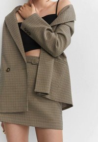 Blazer marrón a cuadros con un corte oversize, top corto negro debajo, y falda a cuadros a juego. Botones visibles y diseño entallado.