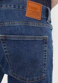 Jeans i denim med en medelblå tvätt, med en läderlapp på midjan prydd med "MTWTFSS WEEKDAY" och vanliga bakfickor.