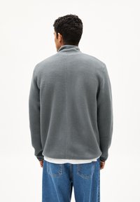 Giacca in fleece grigia con colletto alto, caratterizzata da una superficie testurizzata e polsini a coste, indossata sopra una camicia bianca e jeans blu.