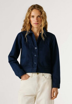 Mujer con cabello rubio ondulado que lleva una chaqueta azul marino abotonada y pantalones beige claro, posando contra un fondo liso con una mano en el bolsillo.