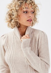 Suéter beige de punto con cuello chal, patrones texturizados, puños acanalados y hombros abullonados. Material suave de peso medio.
