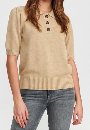 Pull polo en tricot beige avec des manches courtes bouffantes, trois boutons marron, et un ourlet et des poignets côtelés, associé à un jean gris.
