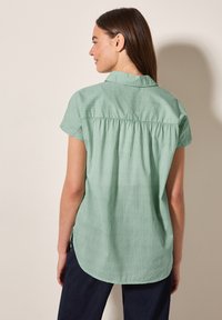 Camicetta verde a maniche corte con colletto, caratterizzata da righe verticali e dettaglio arricciato sul retro. Tessuto morbido e leggero.