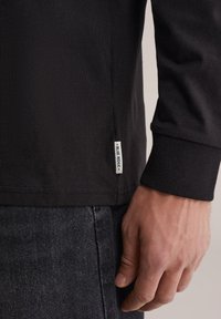 Juoda ilgomis rankovėmis marškinėliai su gofruotais rankogaliais, šonine siūle esančia etikete su užrašu „BLUE RIDGE“ ir lygiu, tekstūruotu audinio paviršiumi.