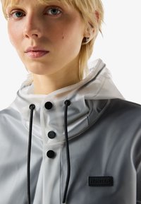 Nahaufnahme einer Person, die einen durchsichtigen grauen Regenmantel mit schwarzen Knöpfen und Kordelzügen trägt, mit einem sichtbaren "HUNTER"-Logo-Patch.