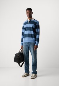 Homme portant un polo à rayures bleues à manches longues, un jean et des baskets, tenant un sac de sport en cuir noir, se tenant devant un fond blanc uni.