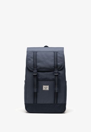 Marinfärgad tygryggsäck med klaffstängning, två justerbara remmar, framficka och rektangulär form. Har vit sömnad och en logotypapplikation.